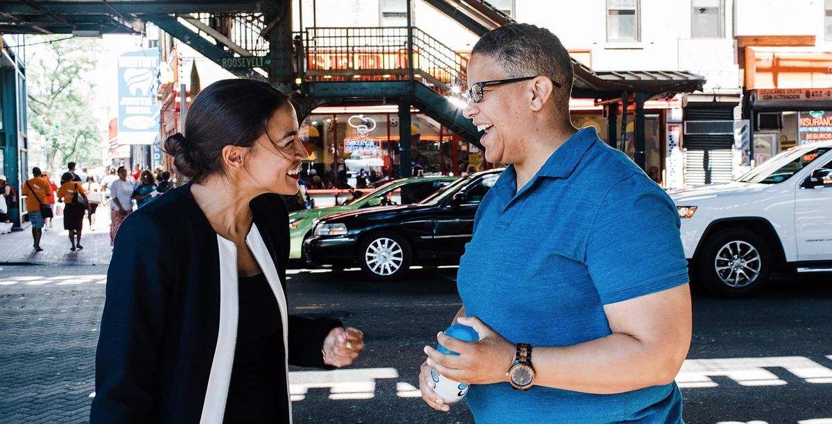 Alexandria Ocasio-Cortez, wearing a dark blazer, converses with Kerri Evelyn Harris, clad in a blue polo, on a vibrant city street. They are both smiling, engaged in a lively discussion. In the background, the steel beams of an elevated train line loom above amidst the urban bustle, with pedestrians and storefronts lining the sunlit sidewalk.